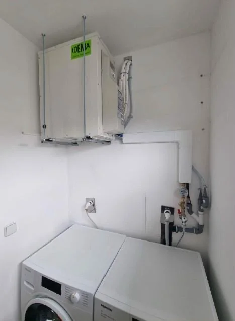 Deux modules intérieurs de climatisation installés en hauteur contre le mur d'une buanderie, au-dessus d'un lave-linge, raccordés par des gaines blanches.